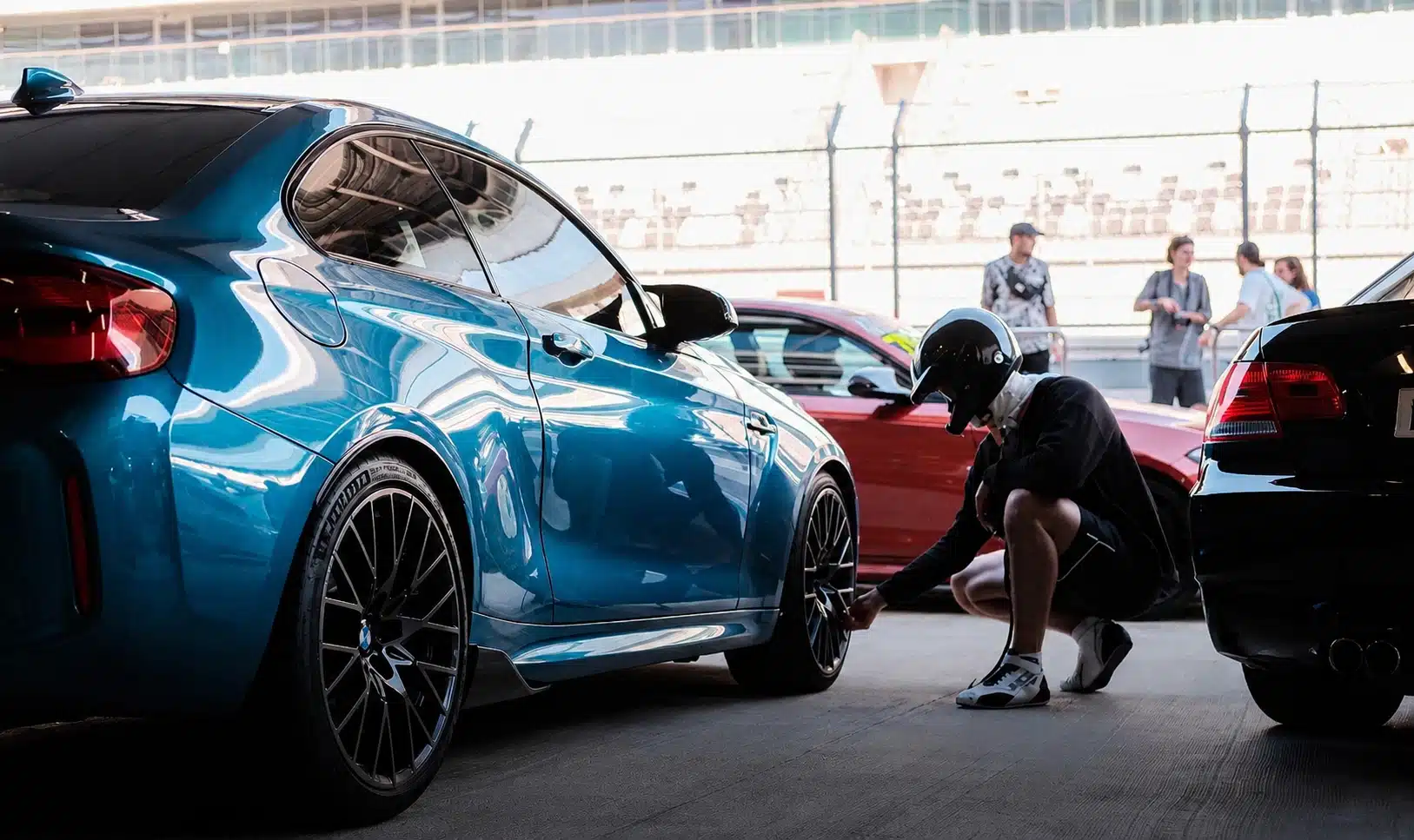 Ein Schüler mit Helm hockt sich hin und überprüft das Rad eines blauen BMW M2 in der Box, im Hintergrund sind weitere Rennwagen und Zuschauer an der Streckenabsperrung zu sehen.