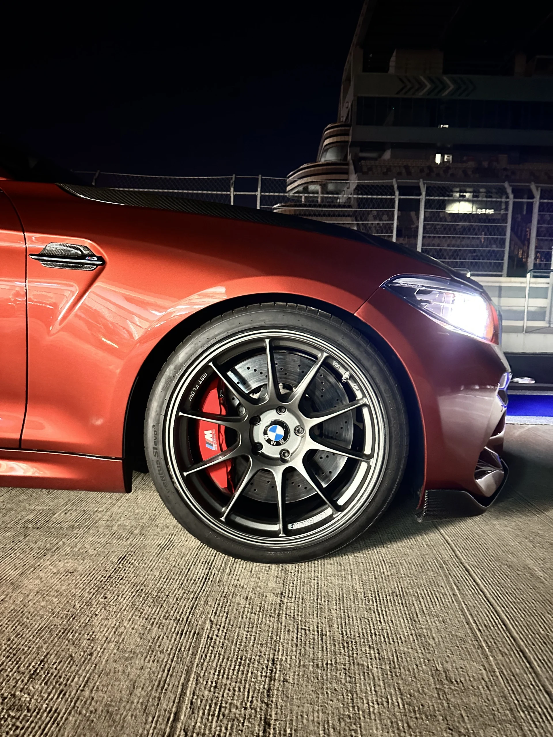 Nahaufnahme des Vorderrads eines orangefarbenen BMW M mit rotem M Performance-Bremssattel, gelochter Bremsscheibe und Carbon-Splitter, aufgenommen nachts auf der Autobahn bei eingeschaltetem Scheinwerfer.