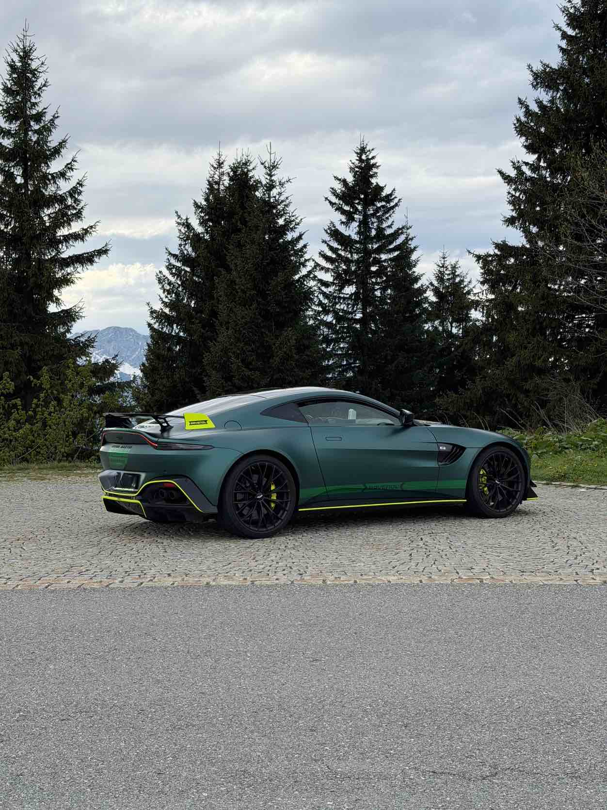 Aston Martin Vantage F1 Safety Car in mattem Brit-Green, Rückansicht auf einem gepflasterten Parkplatz, umgeben von Tannen und Berggipfeln im Hintergrund.