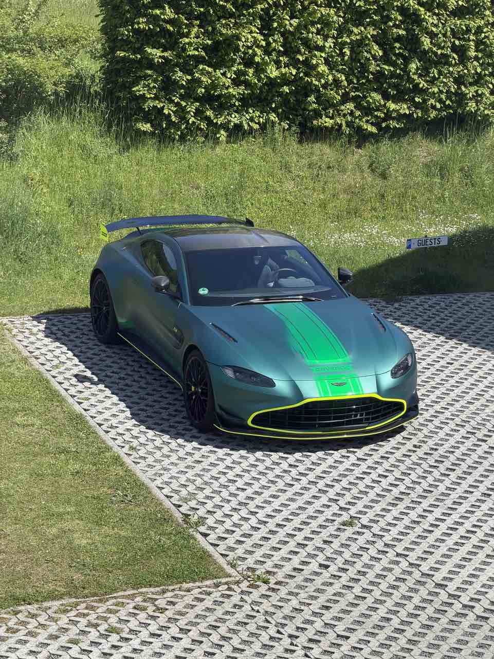 Ein Aston Martin Vantage F1 Safety Car in mattem British Green auf einem Parkplatz mit Gitterfliesen, umgeben von Grün, leicht von oben aufgenommen.
