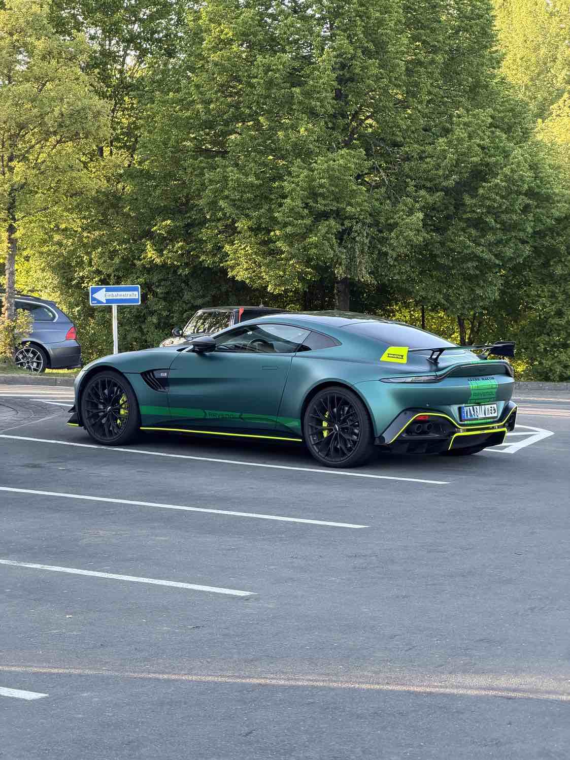 Aston Martin Vantage F1 Safety Car in mattem britischem Grün auf einem Parkplatz in Deutschland, Seitenansicht, grüne Bäume im Hintergrund.