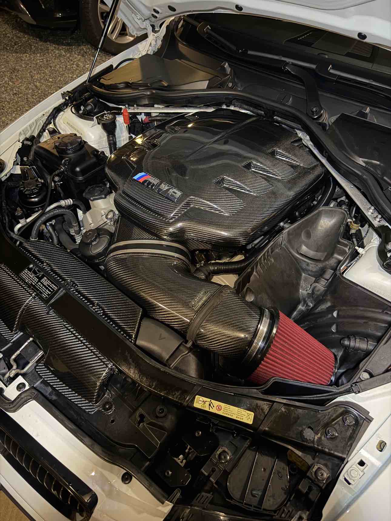Der Motorraum eines weißen BMW M3 E92 mit V8-Motor und einem Eventuri-Ansaugsystem aus Carbon, einem roten Sportluftfilter und einer Motorabdeckung aus Carbon mit M-Streifen.