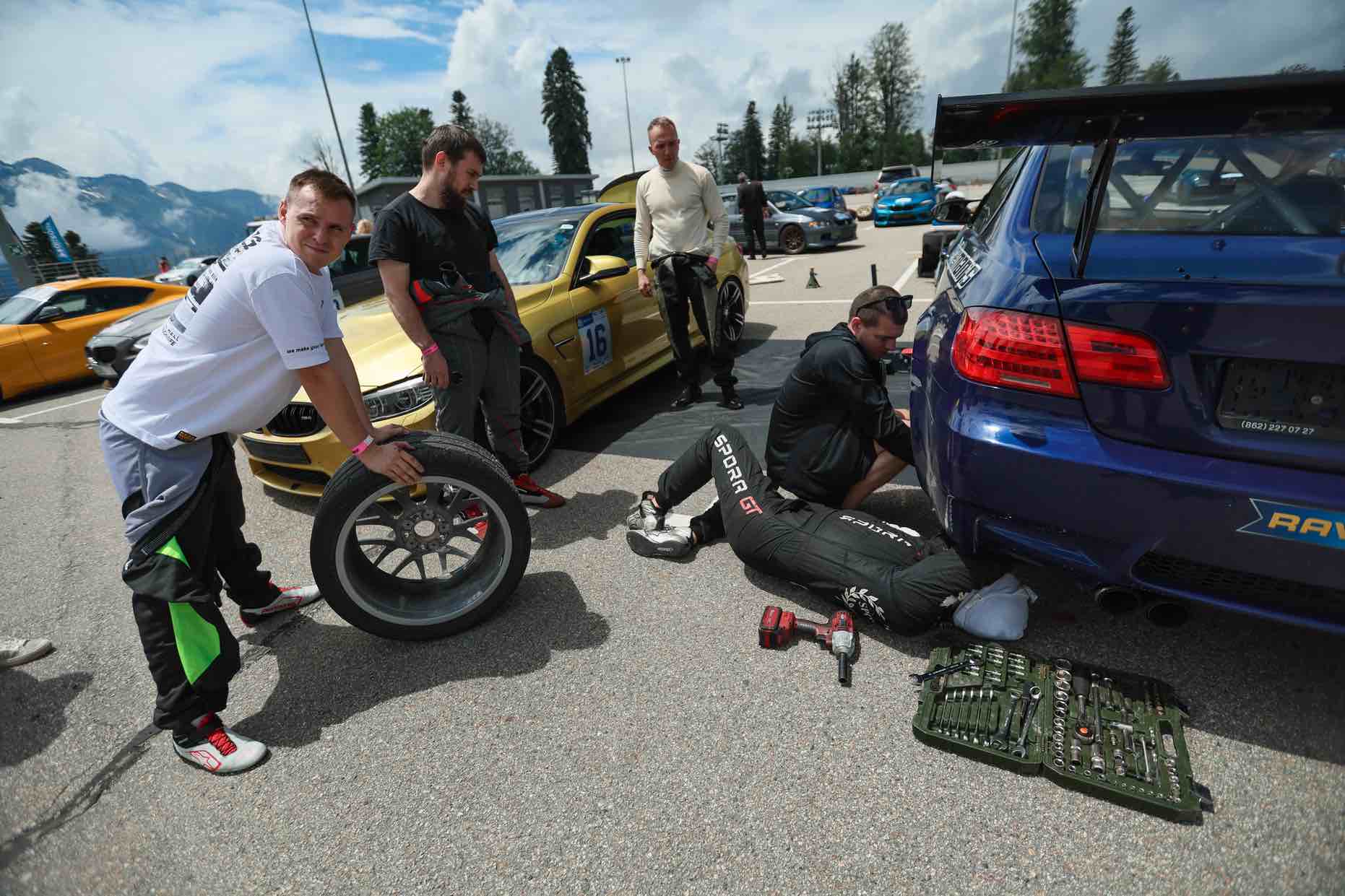 Mechaniker wechseln die Reifen an einem blauen BMW M3 im Fahrerlager des Gazprom-Bergrennens 2026, im Hintergrund ein goldener BMW mit der Startnummer 16 und eine Berglandschaft.