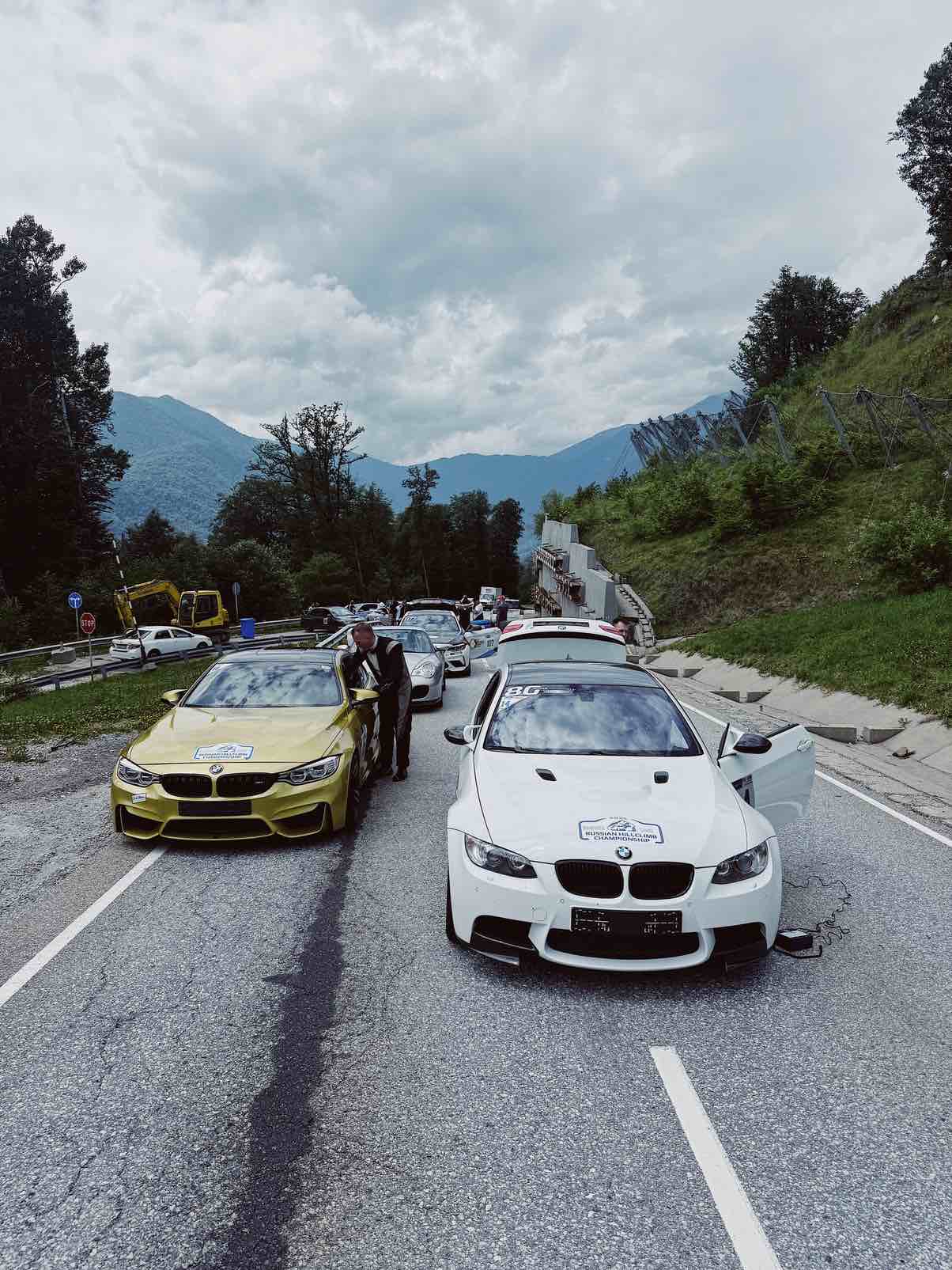 Die Startaufstellung bei der „Russian Hillclimb Championship Gazprom“ in den Bergen: Ein gelber BMW M4 und ein weißer BMW M3 E92 stehen nebeneinander auf der Bergstrecke, im Hintergrund sind ein bewölkter Himmel und grüne Berghänge zu sehen.
