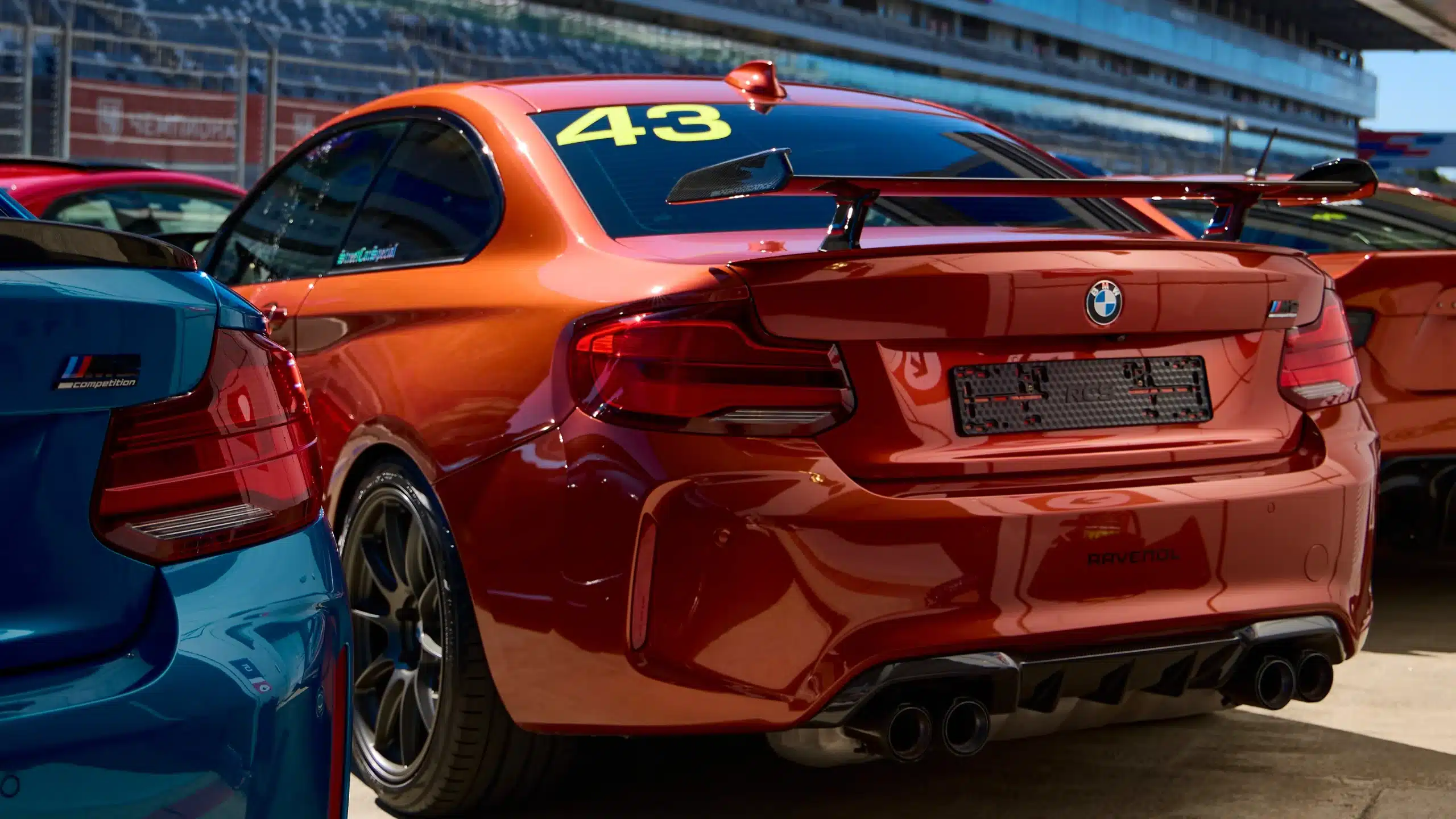 BMW M2 Competition mit der Startnummer 43, Rückansicht in der Boxengasse, daneben ein blauer BMW M2 Competition, im Hintergrund die Rennstrecke.