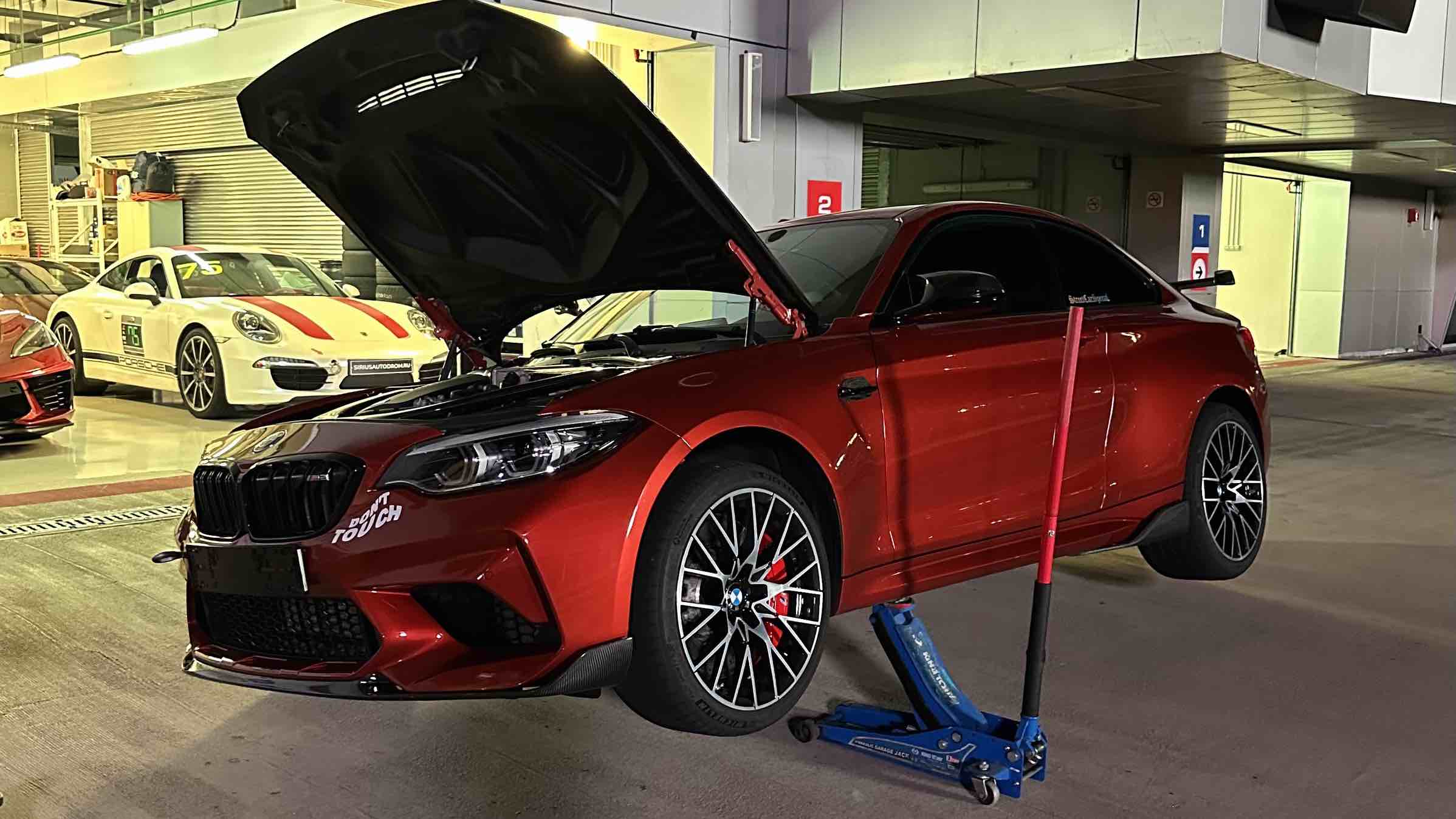 Ein orangefarbener BMW M2 Competition F87 auf einem Wagenheber während der Wartung vor dem Track-Day; im Hintergrund stehen Autos in der Box