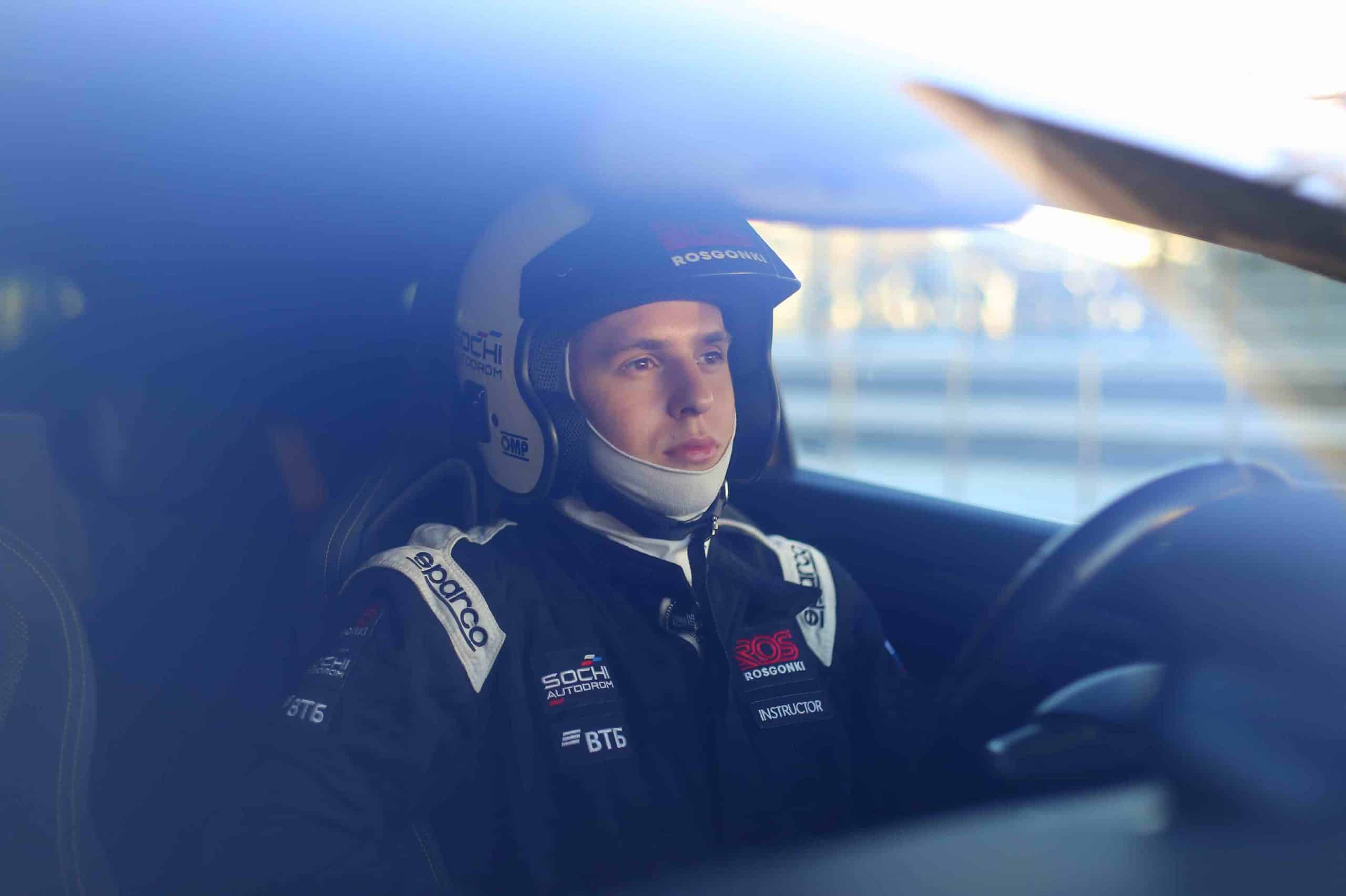 Ich bin Fahrlehrer am Sochi Autodrom und sitze vor dem Start und dem Treffen mit dem Kunden im Auto, bekleidet mit einem Overall mit dem Aufnäher „Fahrlehrer“.