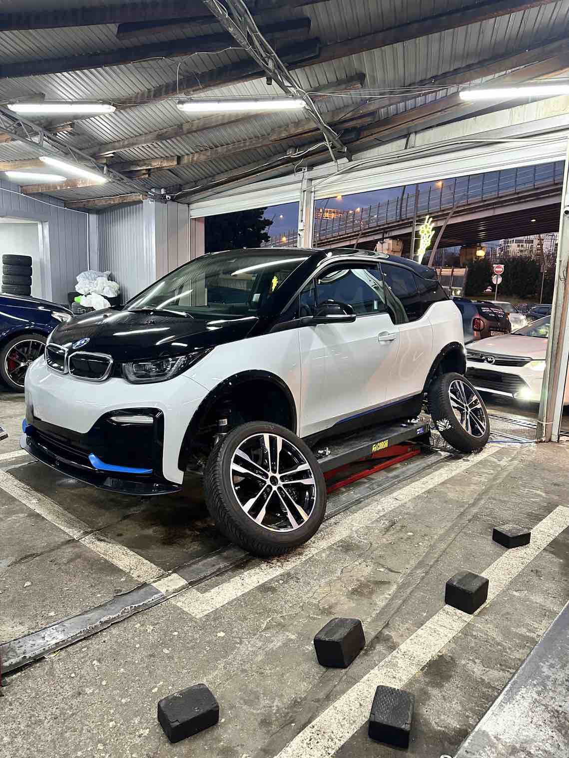 Ein BMW i3s auf der Hebebühne, dessen Räder abmontiert sind, die aber neben dem Auto liegen